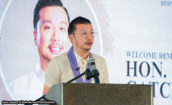 Man with glasses at a podium giving a speech, microphone in front, with a large portrait and 'Welcome' banner in the background.